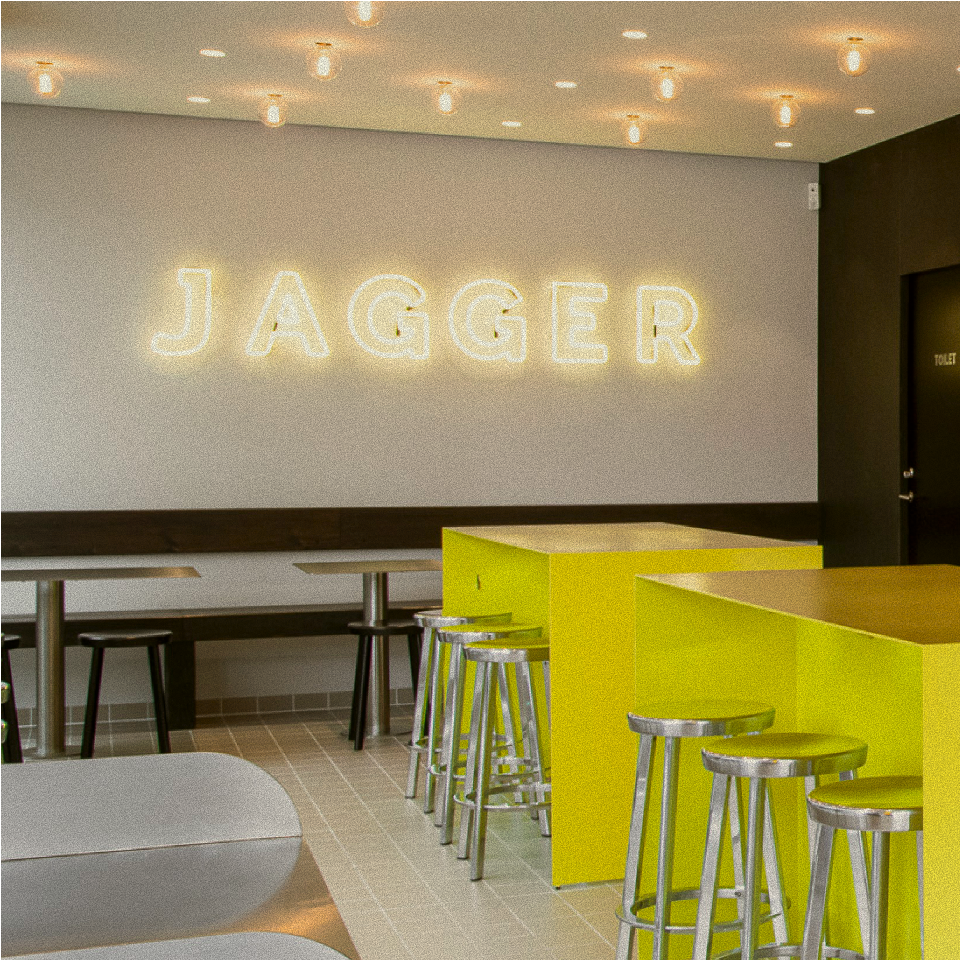 Interior of a Jagger restaurant with a neon Jagger sign, yellow high tables and steel bar stools